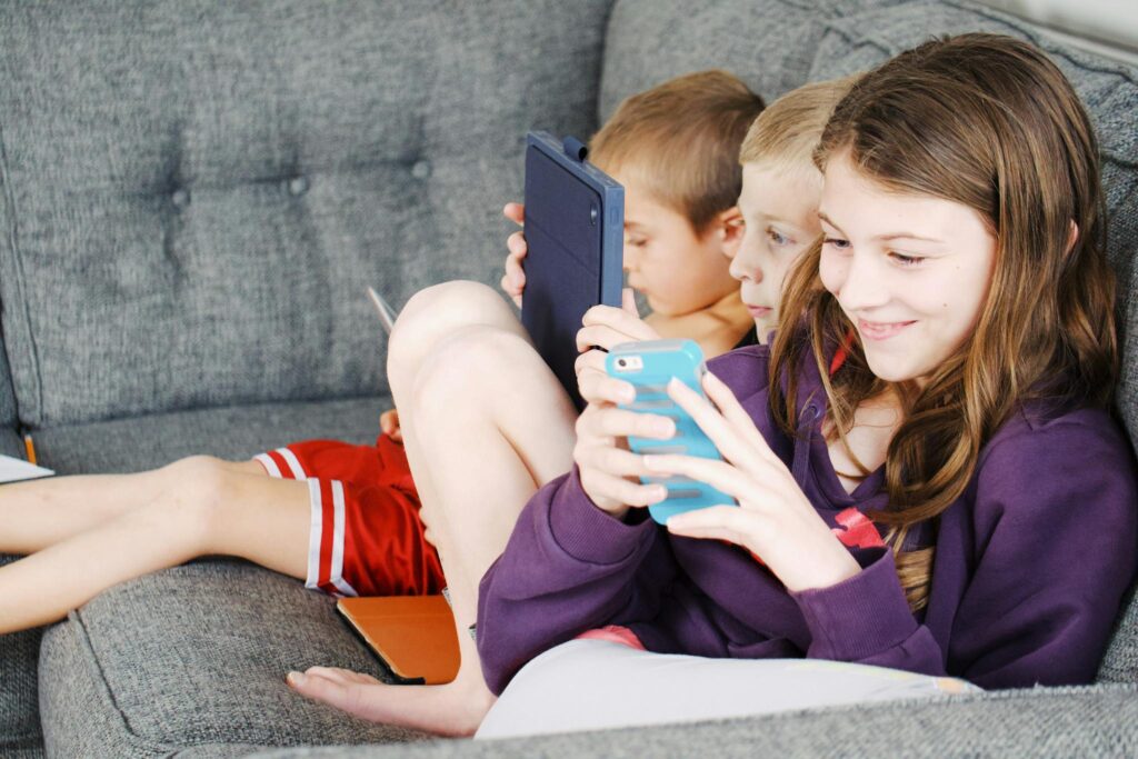 Three children sitting on a couch, engaged with electronic devices, smiling and enjoying leisure time.