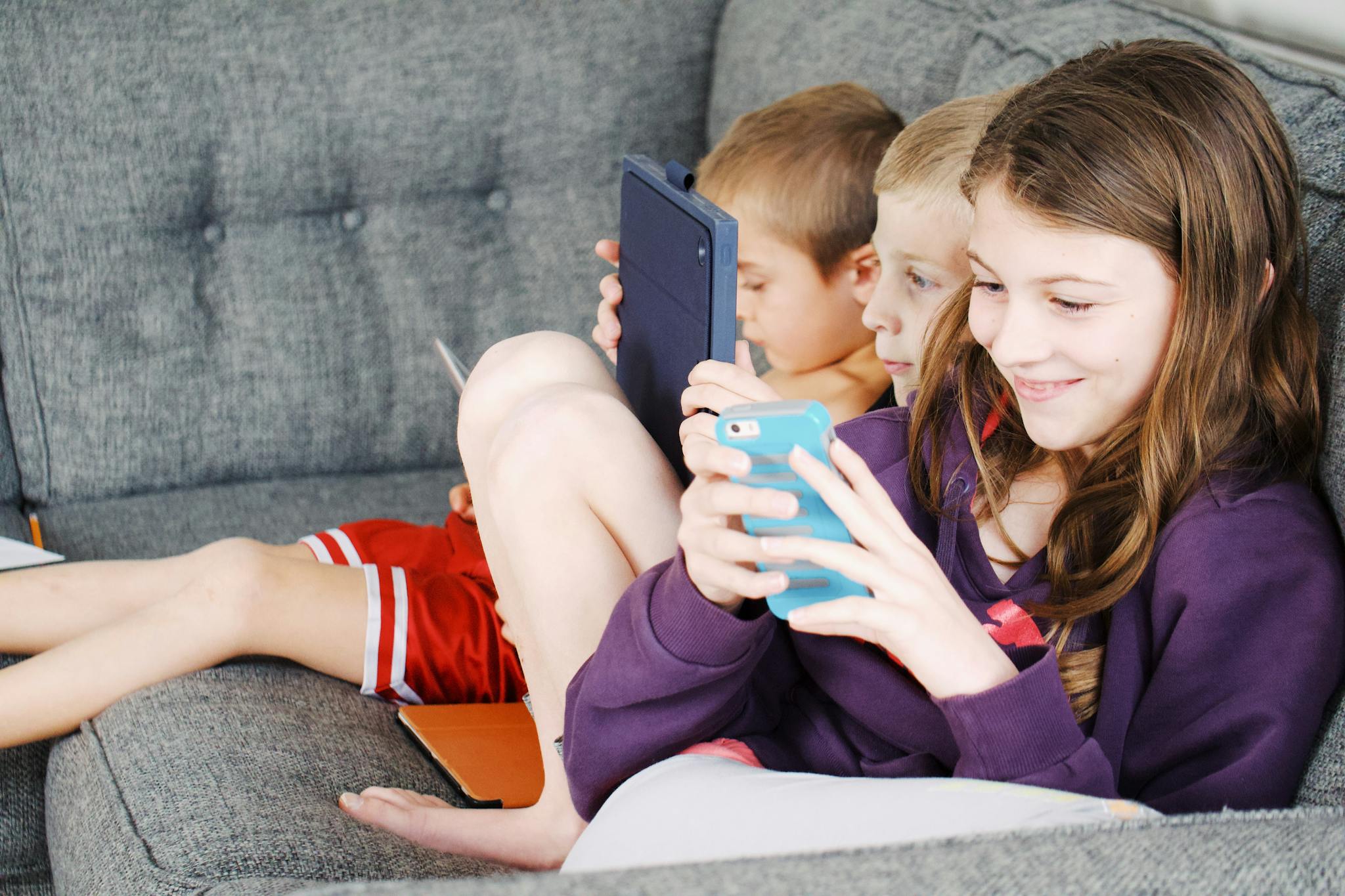 Three children sitting on a couch, engaged with electronic devices, smiling and enjoying leisure time.